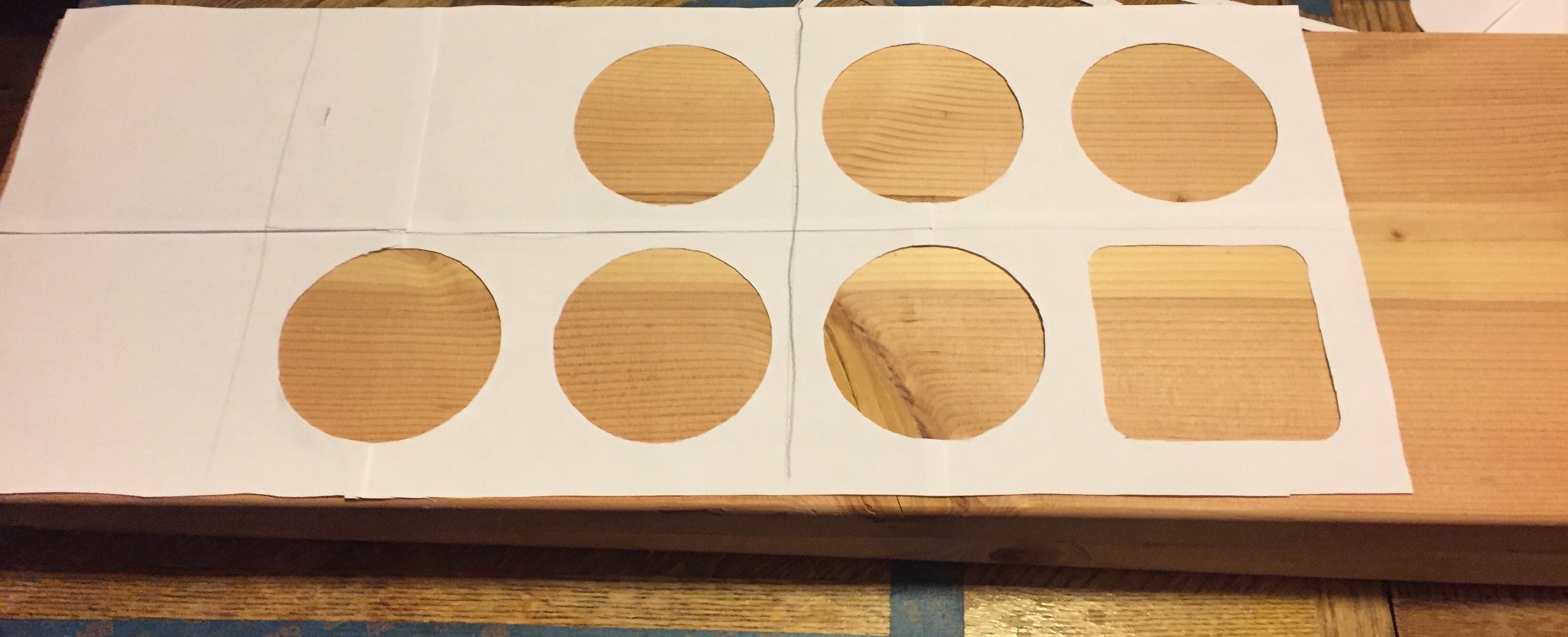 Paper with multiple circular holes and one square hole laid on top of 2×12 board